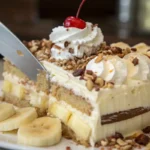 Layered banana split cake with graham cracker crust, vanilla, chocolate, and strawberry ice cream, topped with whipped cream, chocolate syrup, chopped nuts, and maraschino cherries.